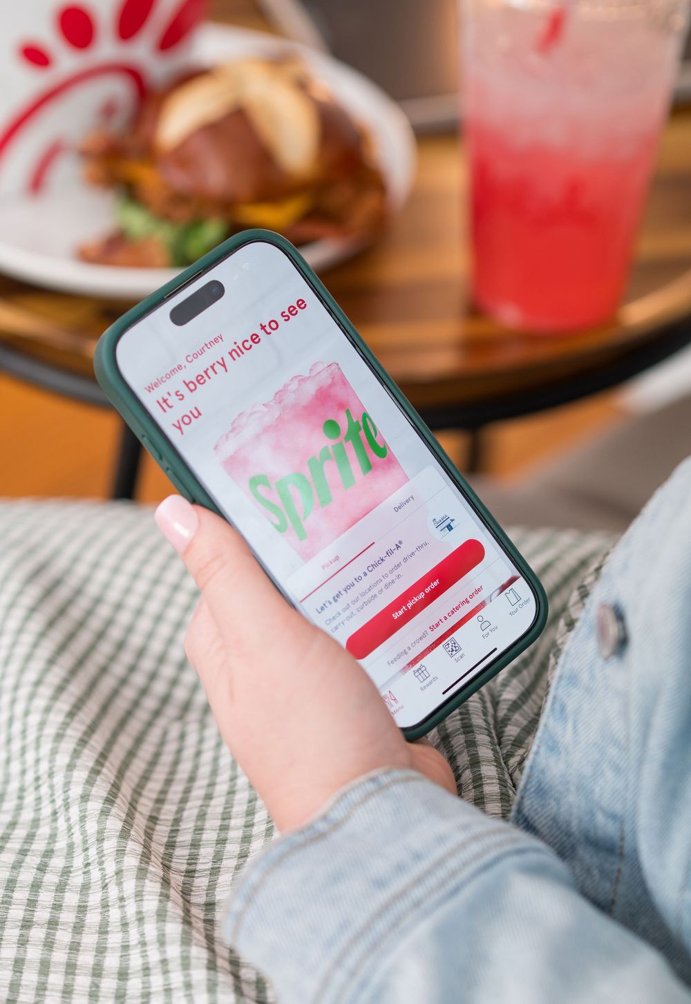 Hand holding phone displaying Chick-fil-A app with food and pink drink in background.