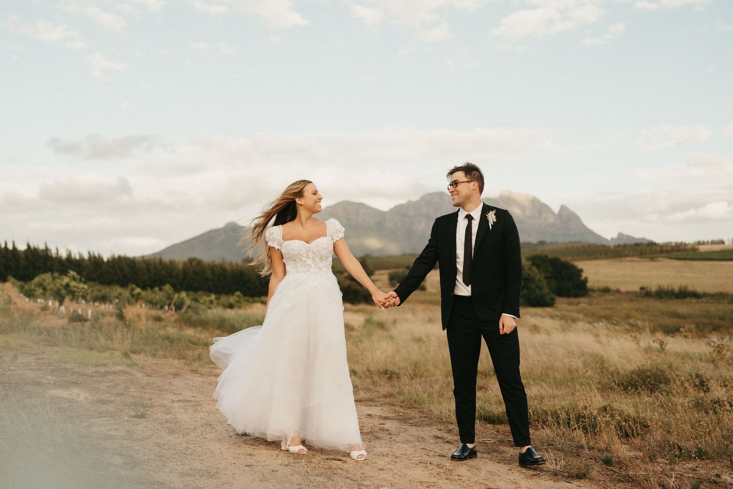 A wedding couple walks along a lakeside path with mountains in the background during golden hour.