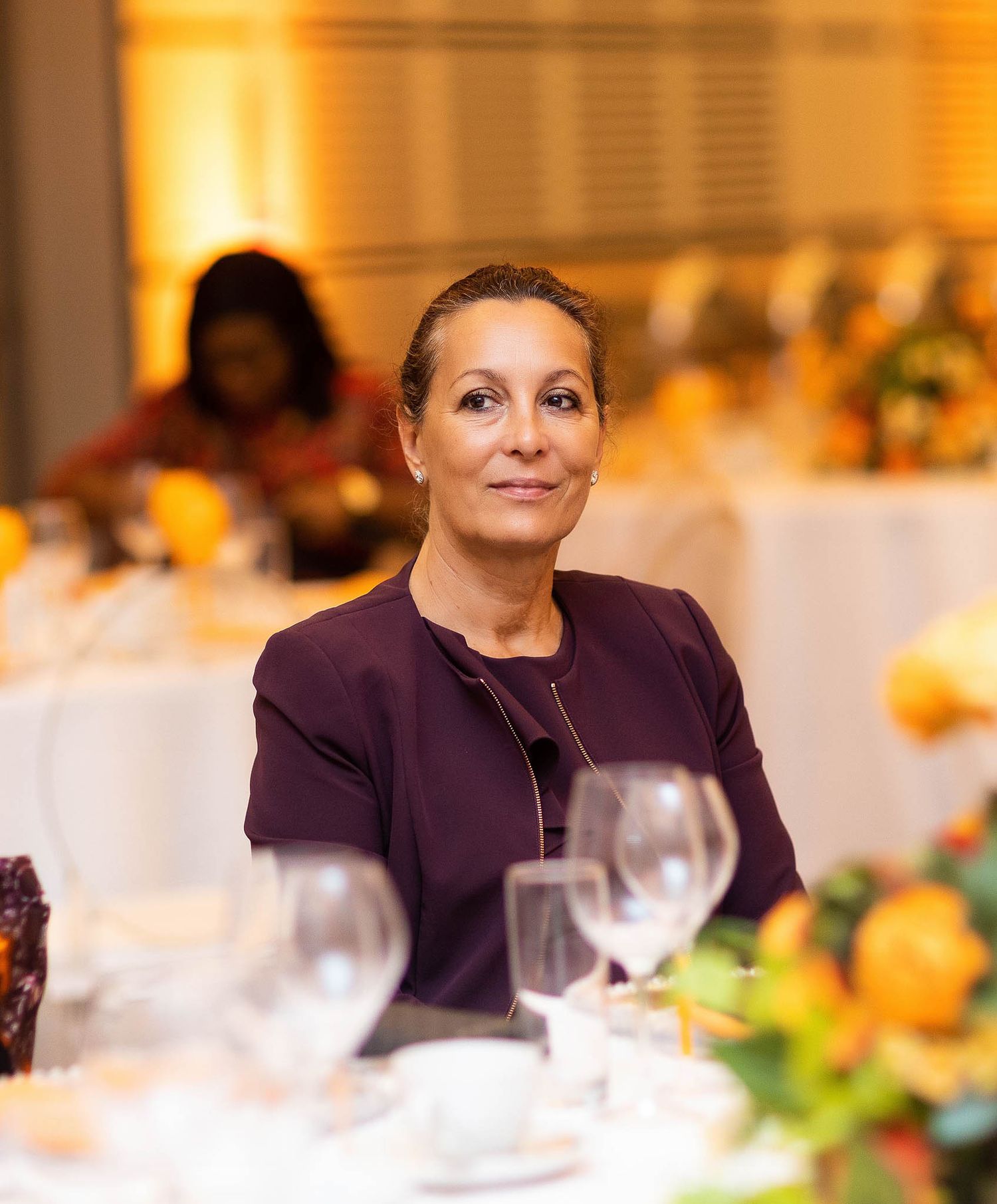 Woman in purple blazer sits at formal dining table with wine glasses and place settings.