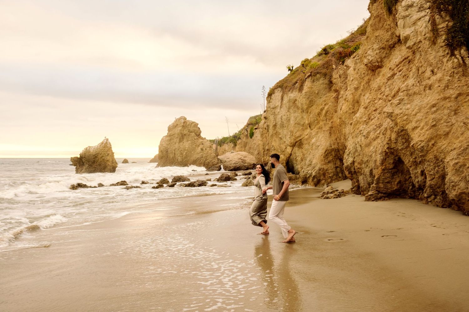 A scenic seascape with rocky cliffs along the shoreline as waves crash on a sandy beach at sunset.
