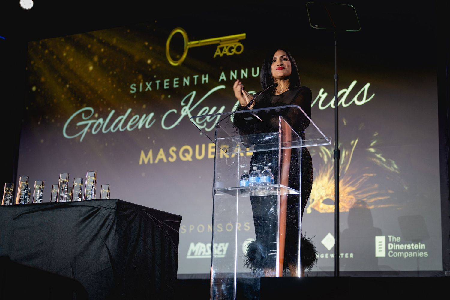 A person stands at a podium on stage during the Golden Keys Masquerade event with a glowing cityscape backdrop.