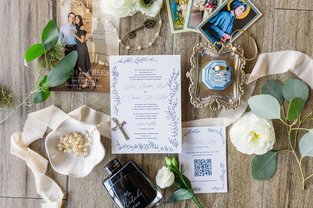 Wedding invitation with white floral accents and eucalyptus leaves laid out on a wooden surface with photos and rings.