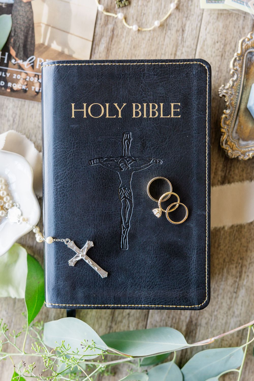 Black leather Holy Bible with golden text and wedding rings against rustic wooden background.