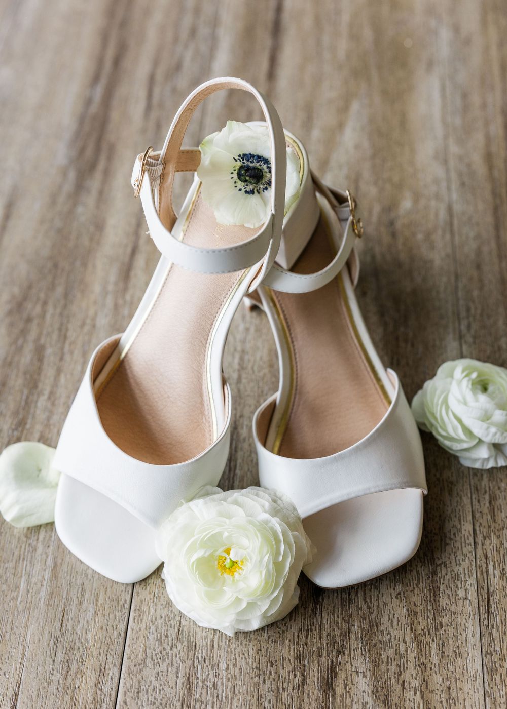 White bridal shoes with delicate straps displayed with white roses on wooden surface.