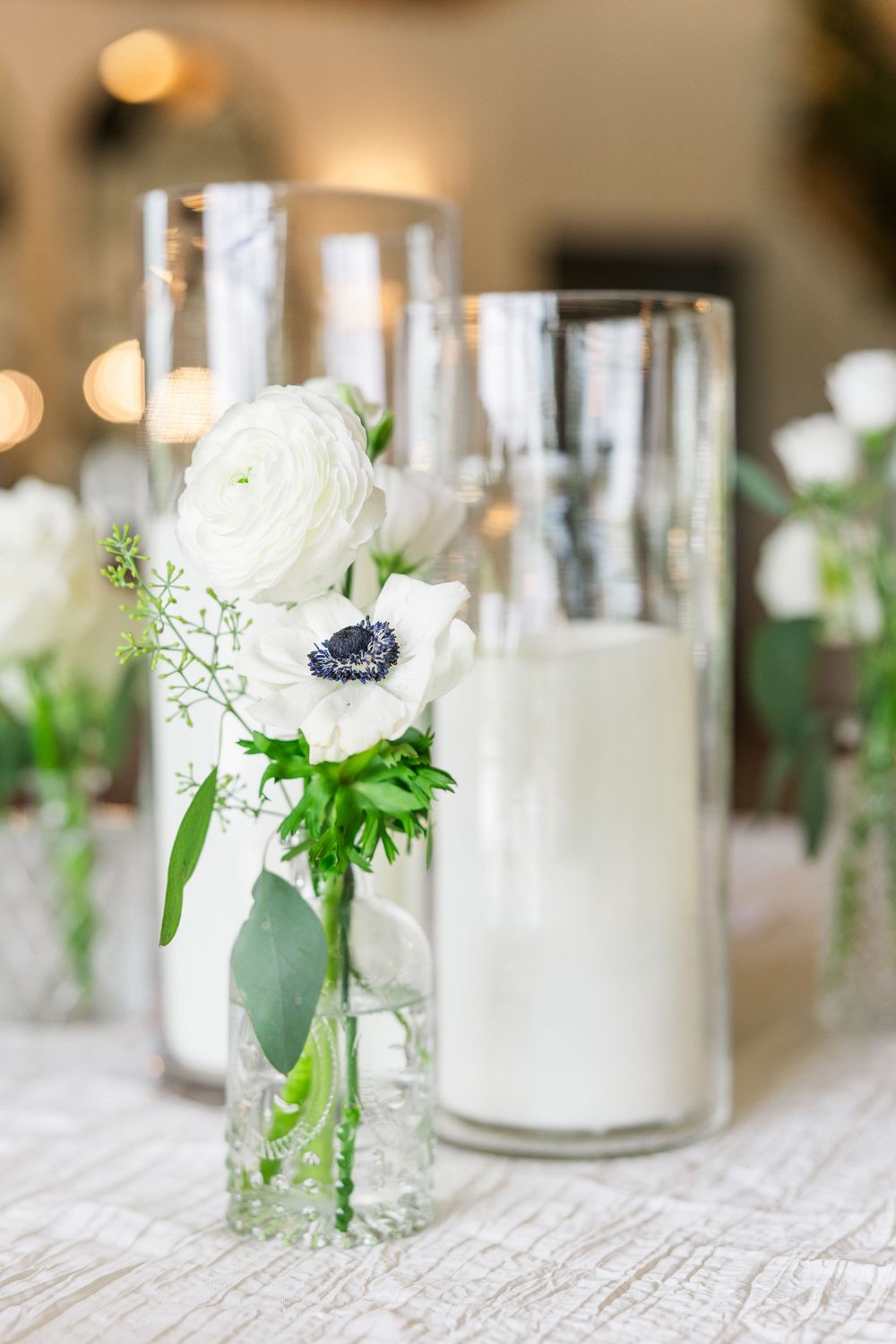 Elegant wedding table decor with white anemone flower and ranunculus in glass vases alongside pillar candles.