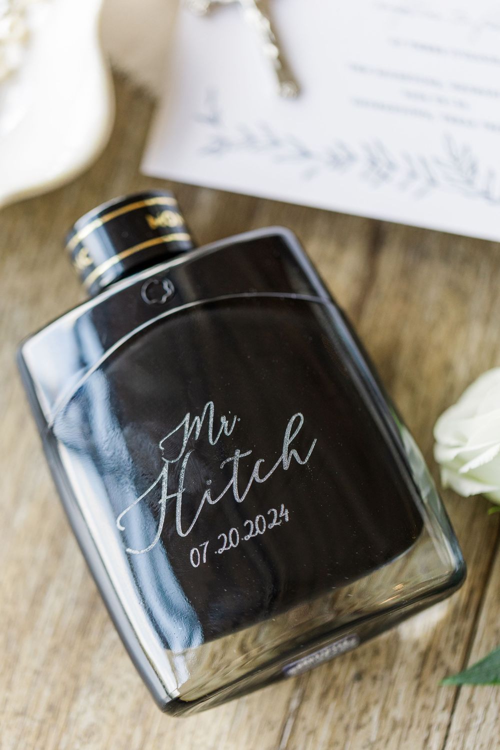 Black metal flask with 'Mr Fletch 01.20.2024' engraved text sits on wooden surface next to white wedding invitation.