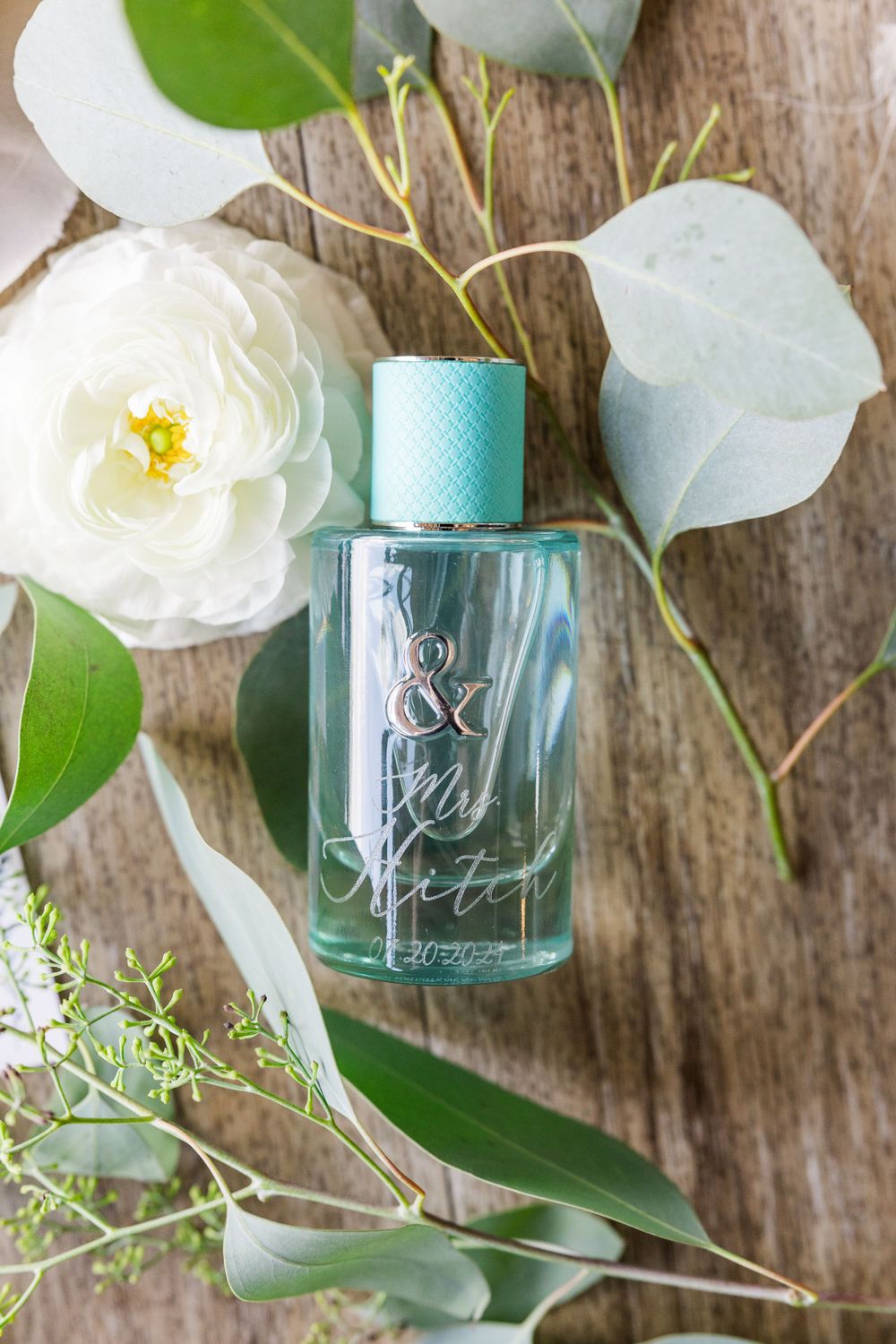 Turquoise perfume bottle with decorative text styled with white flowers and eucalyptus leaves.