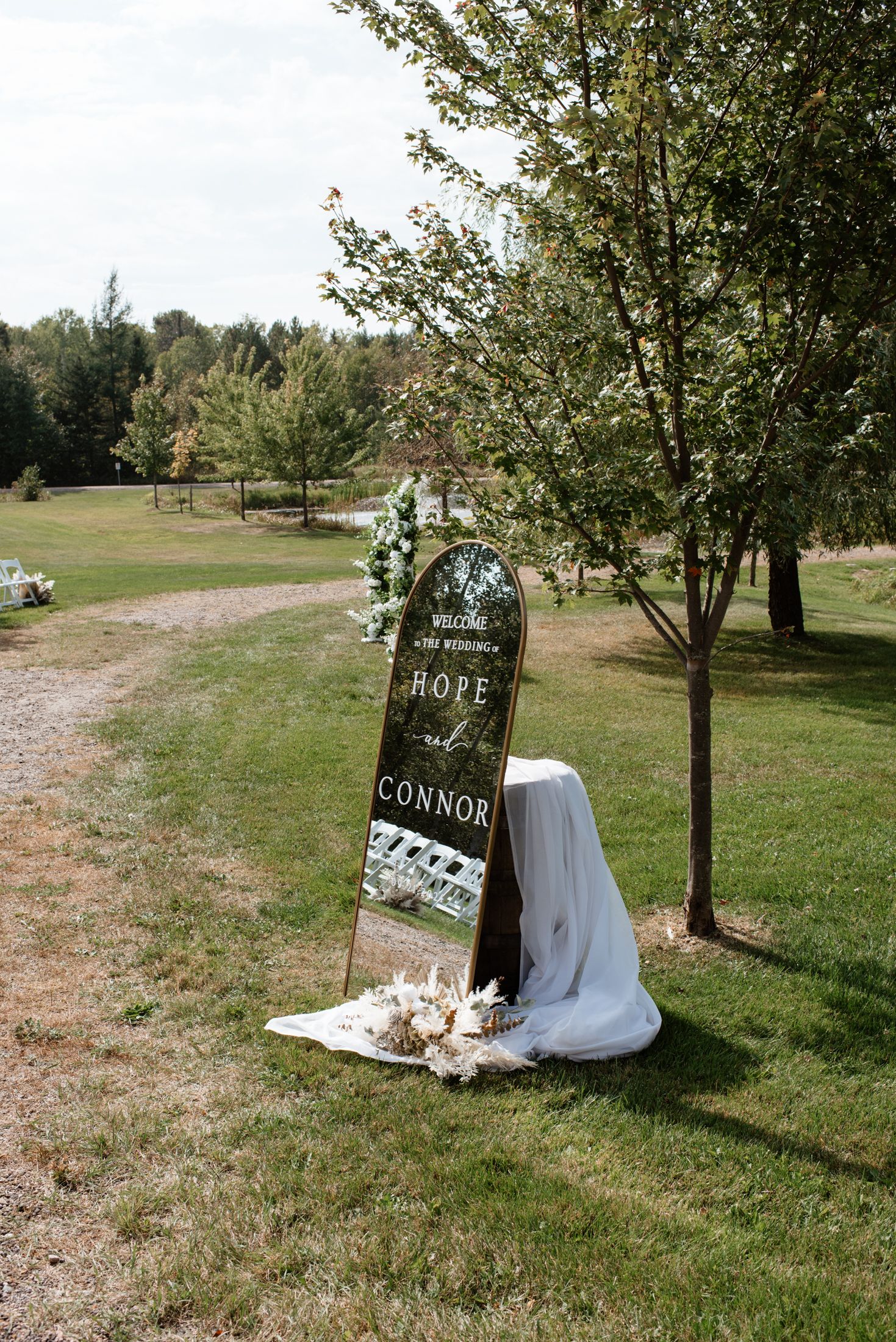 A chalkboard welcome sign propped against a tree at an outdoor wedding venue with white fabric and floral decorations.