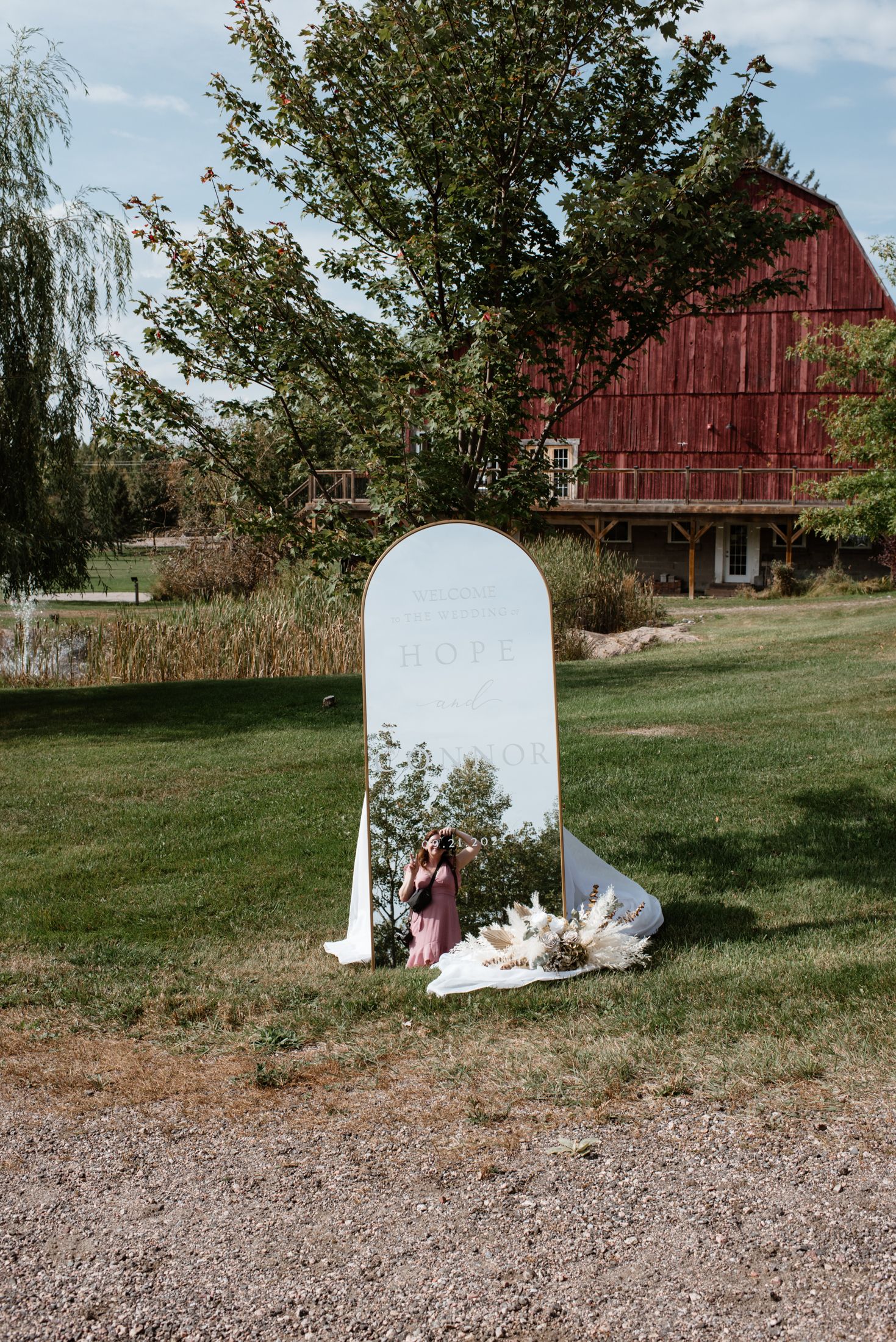 A white freestanding mirror prop sits on grass with a rustic red barn and willow tree in the background.