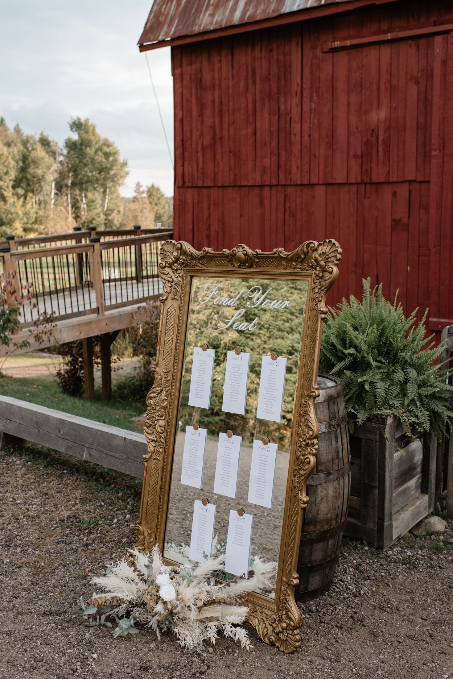 A decorative gold ornate mirror with seating chart display stands outside a rustic red barn with greenery.