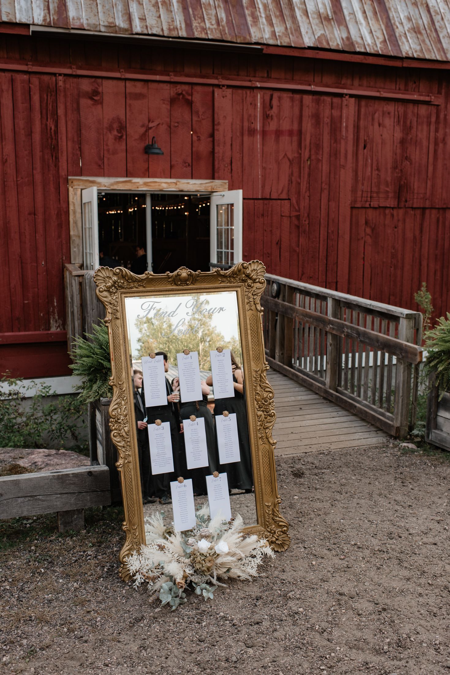 An ornate gold mirror with seating chart cards displayed stands against a rustic red barn wall with a wooden ramp.