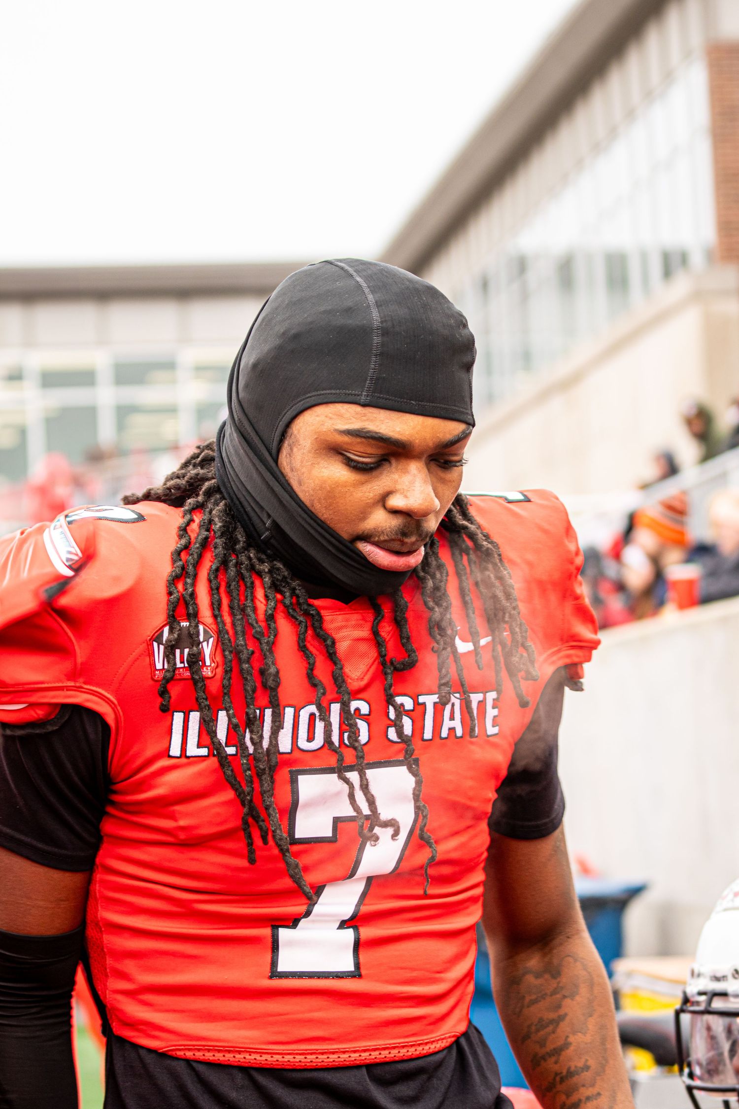 Illinois State football player in red jersey number 7 stands on sidelines wearing black head covering.
