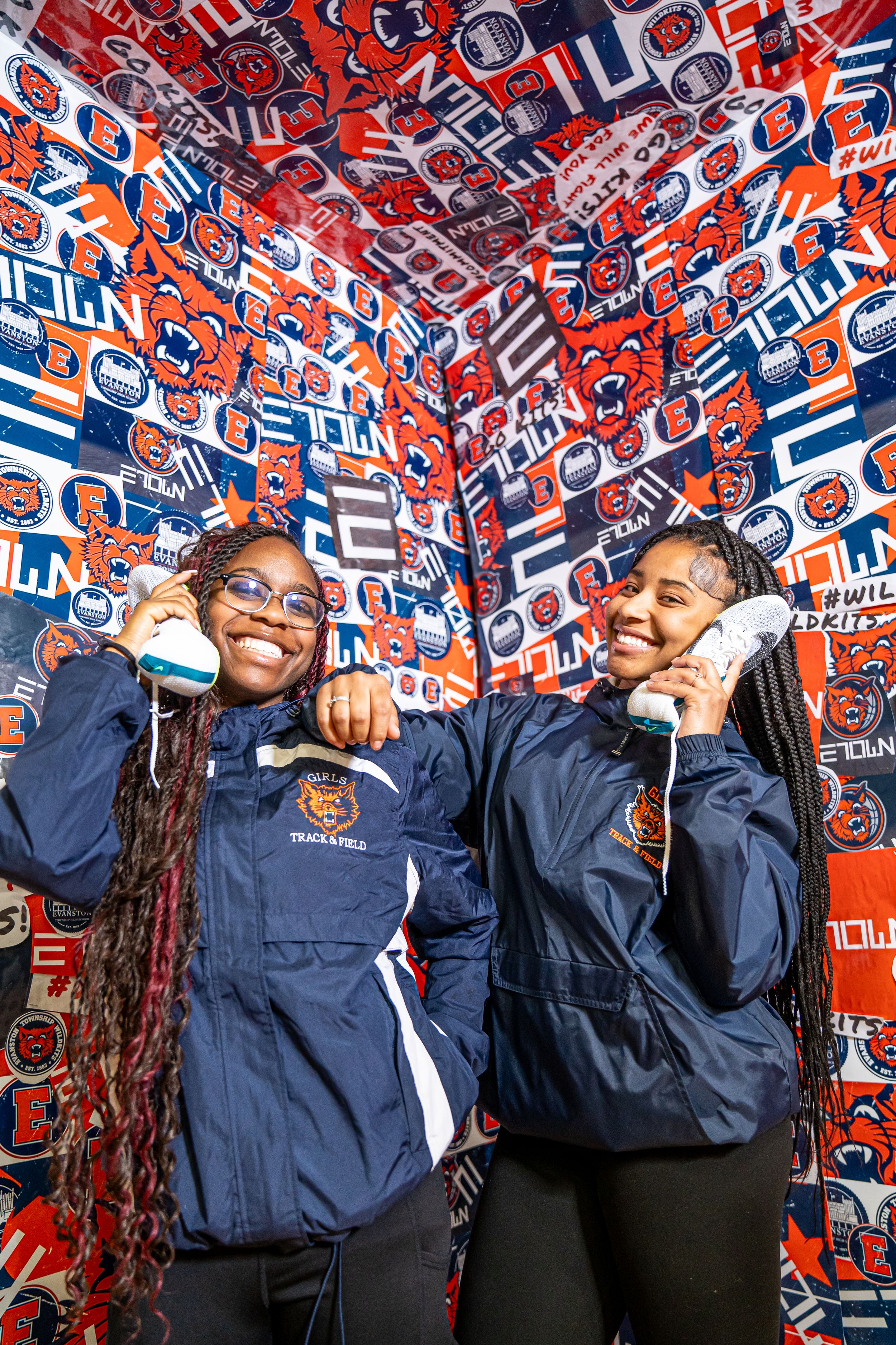 Two basketball fans in navy blue jackets pose against a wall of red and blue team logos and patterns.