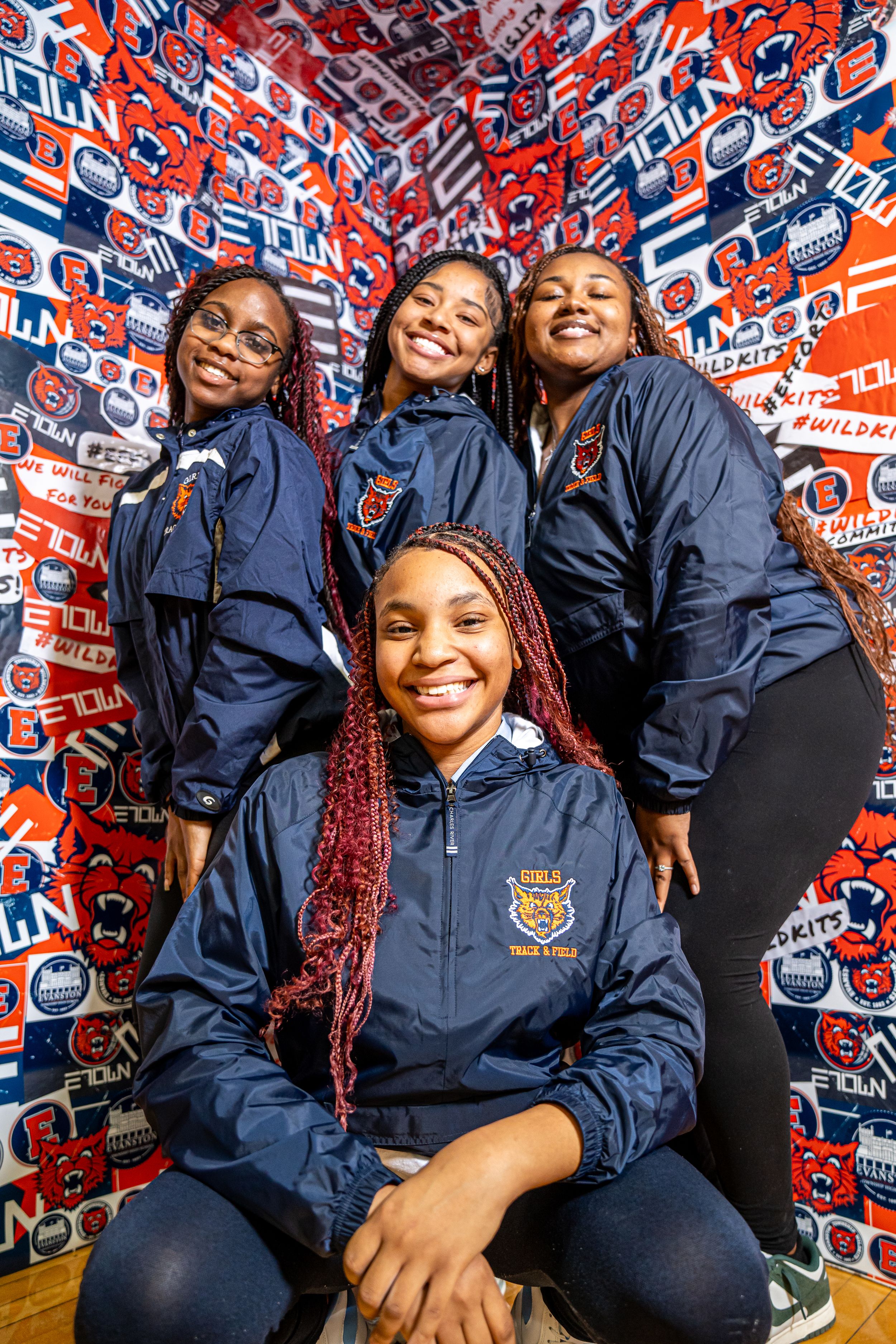People wearing navy blue team jackets pose cheerfully against a wall covered in red and blue sports team logos.