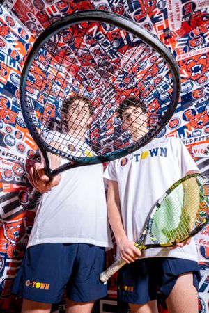 Two tennis players in white shirts and navy shorts hold large rackets against a wall covered in red and blue sports logos.