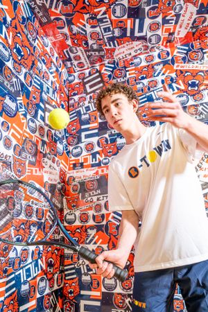 Against a wall covered in orange and blue Gulf Oil logos, someone tosses a tennis ball while looking at the camera.