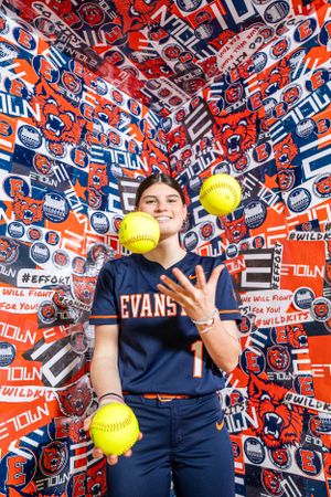 Softball player in navy uniform juggles three yellow softballs against vibrant red and blue patterned background.