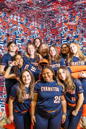 Group wearing navy blue Evanston athletic jerseys poses in front of a wall covered in sports-themed decorations and logos.