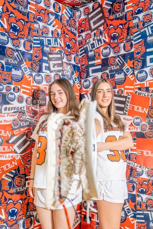 Two people pose in white outfits against a vibrant orange and blue wall covered in sports team logos and graphics.