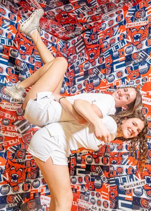 Two people in white outfits pose against a backdrop covered in vintage Pepsi logos and branding elements.