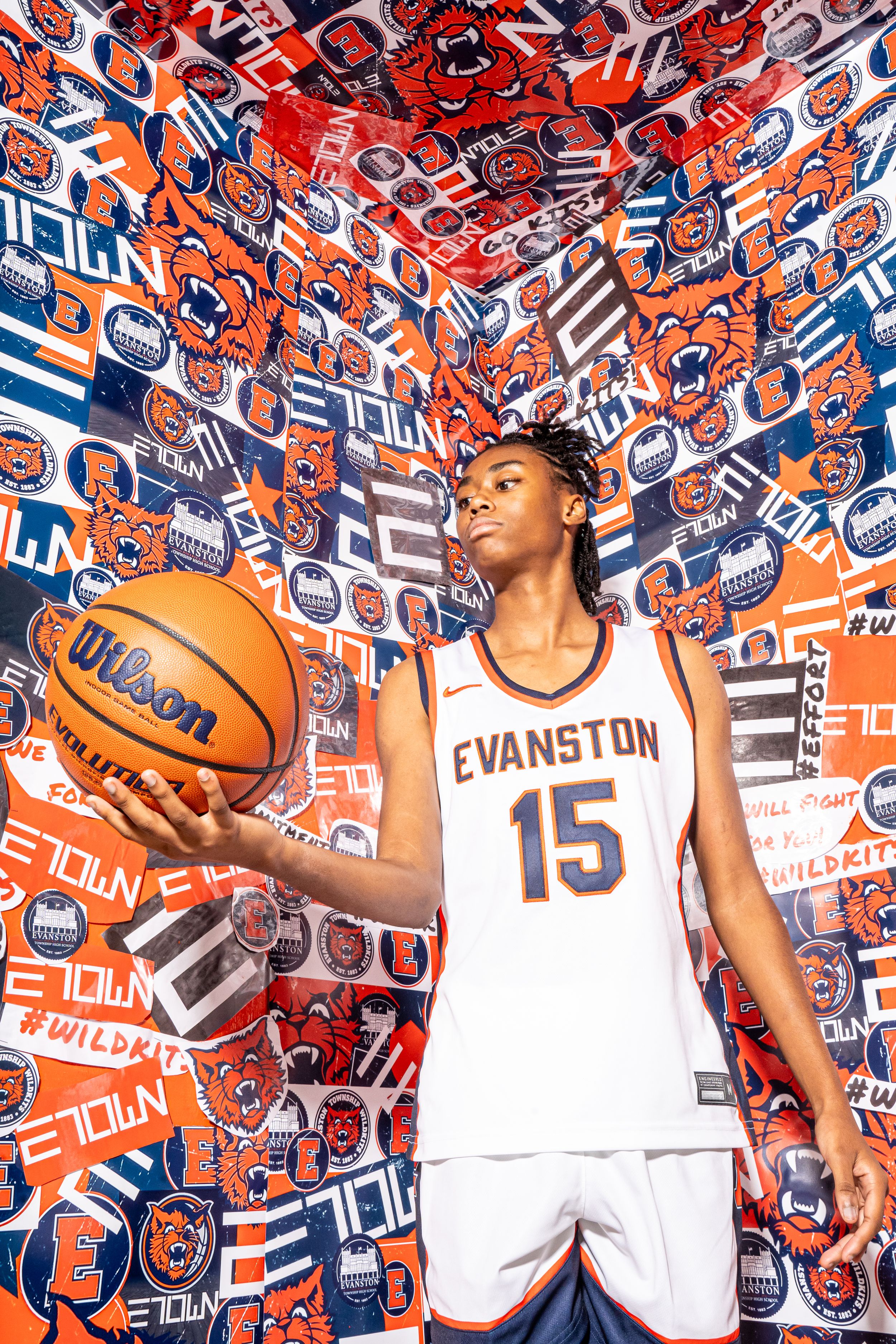 Basketball player in white Evanston #15 jersey poses against vibrant red and blue patterned wall with basketball.