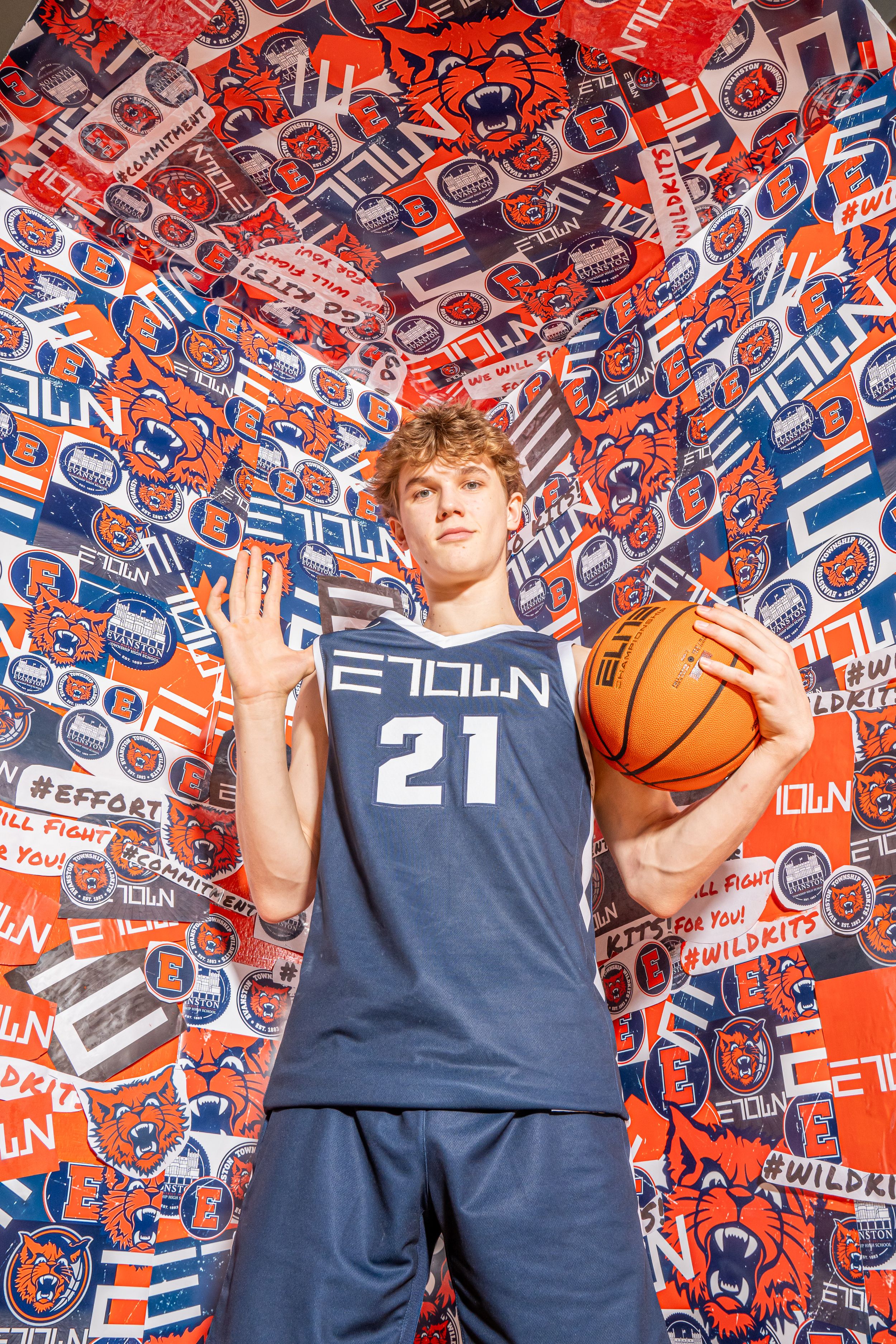 Basketball player in navy blue Filion uniform number 21 stands against vibrant red and orange patterned background.