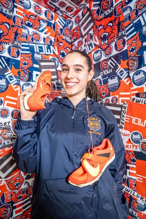 Person in navy blue jacket holds up orange Nike athletic shoes against colorful wall of Edmonton Oilers hockey team logos.