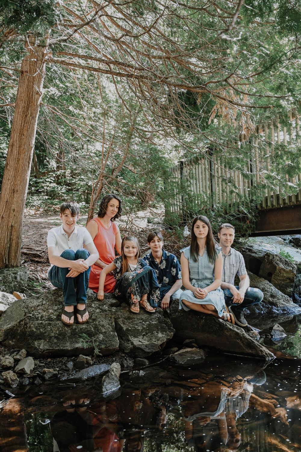 A group sits together on rocks by a peaceful stream in a forest setting with tall trees and dappled sunlight.