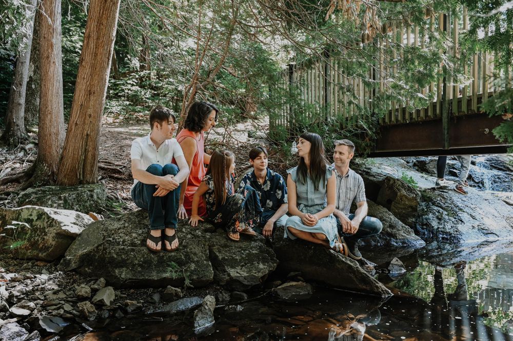 A group sits together on mossy rocks by a flowing stream in a lush forest setting with a wooden bridge in the background.
