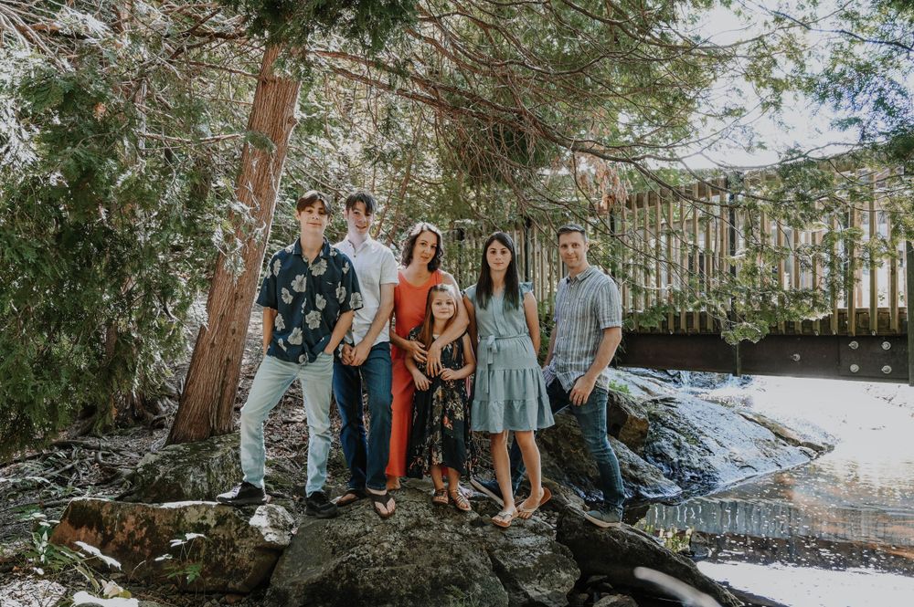Group in casual attire poses together on rocky shoreline surrounded by trees and water in natural outdoor setting.
