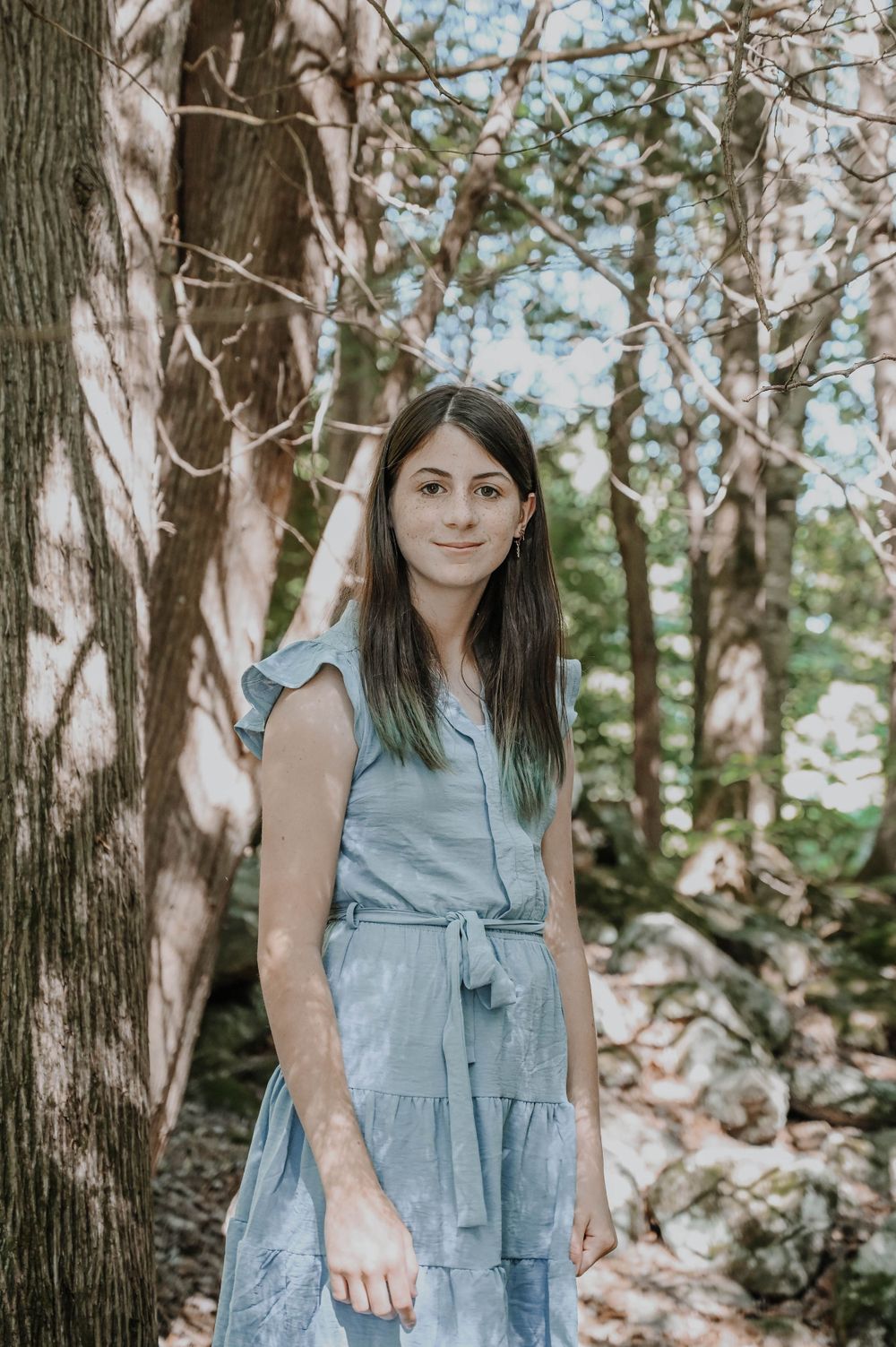 A person in a light blue tiered dress poses against a tree trunk in a serene forest setting.