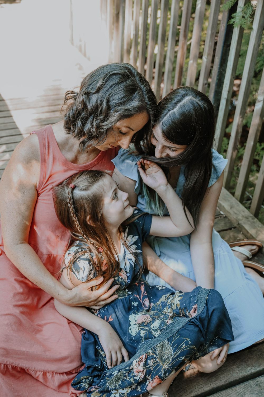 Three people in floral dresses share an intimate family moment together on a wooden deck in soft natural lighting.