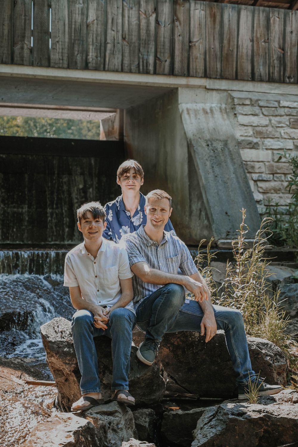 Three people sit on rocks near a small waterfall beneath a rustic wooden bridge surrounded by stone walls.