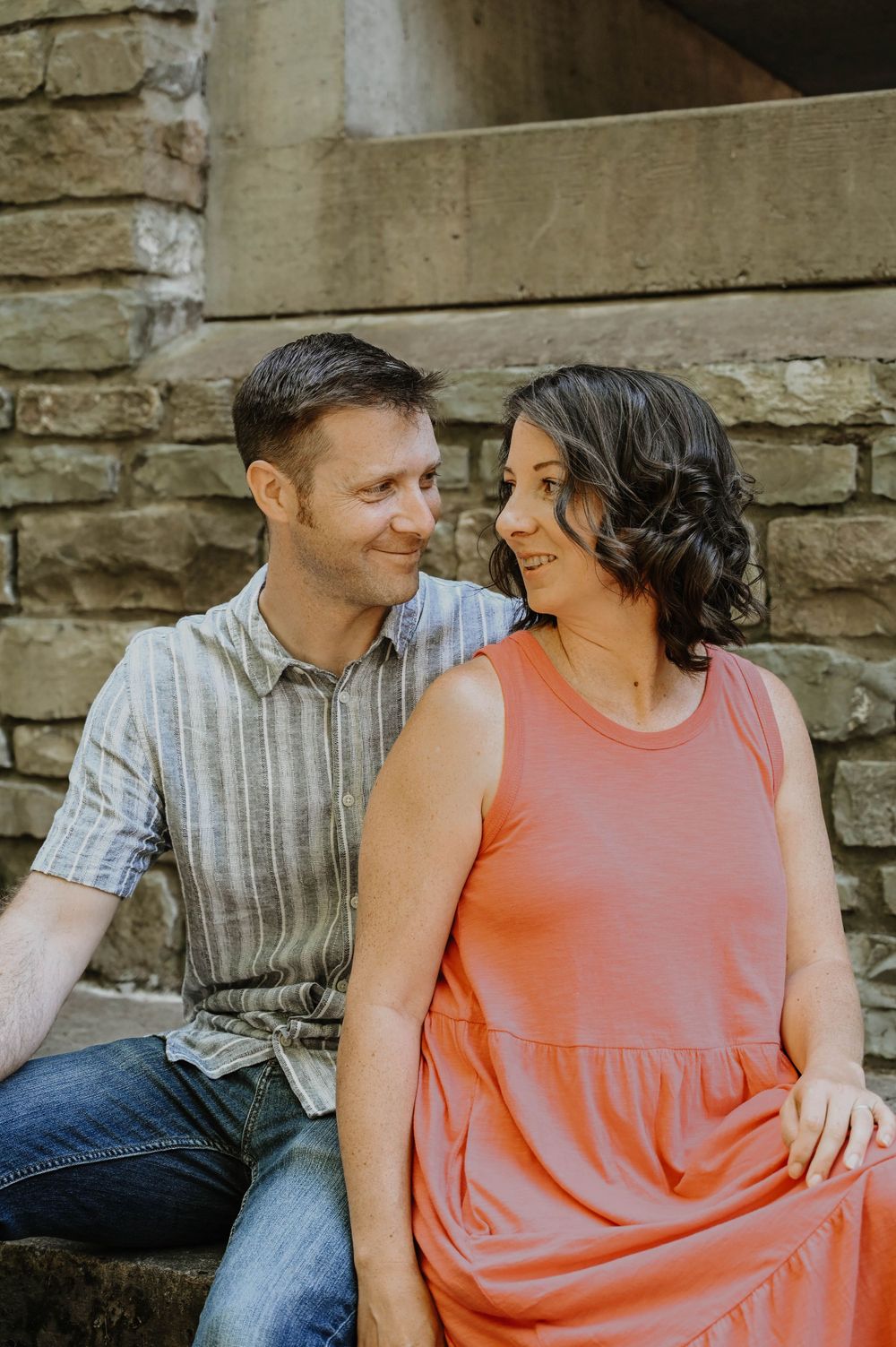 A couple sits together on stone steps during an intimate outdoor photo session wearing casual summer attire.