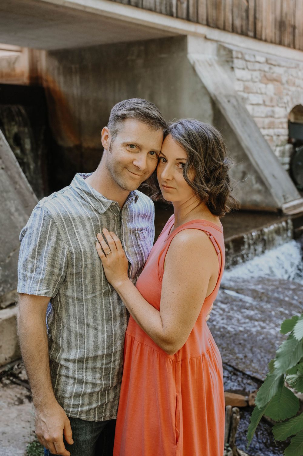A couple poses together outdoors in casual attire, with a coral dress and plaid shirt, against a rustic stone wall background.