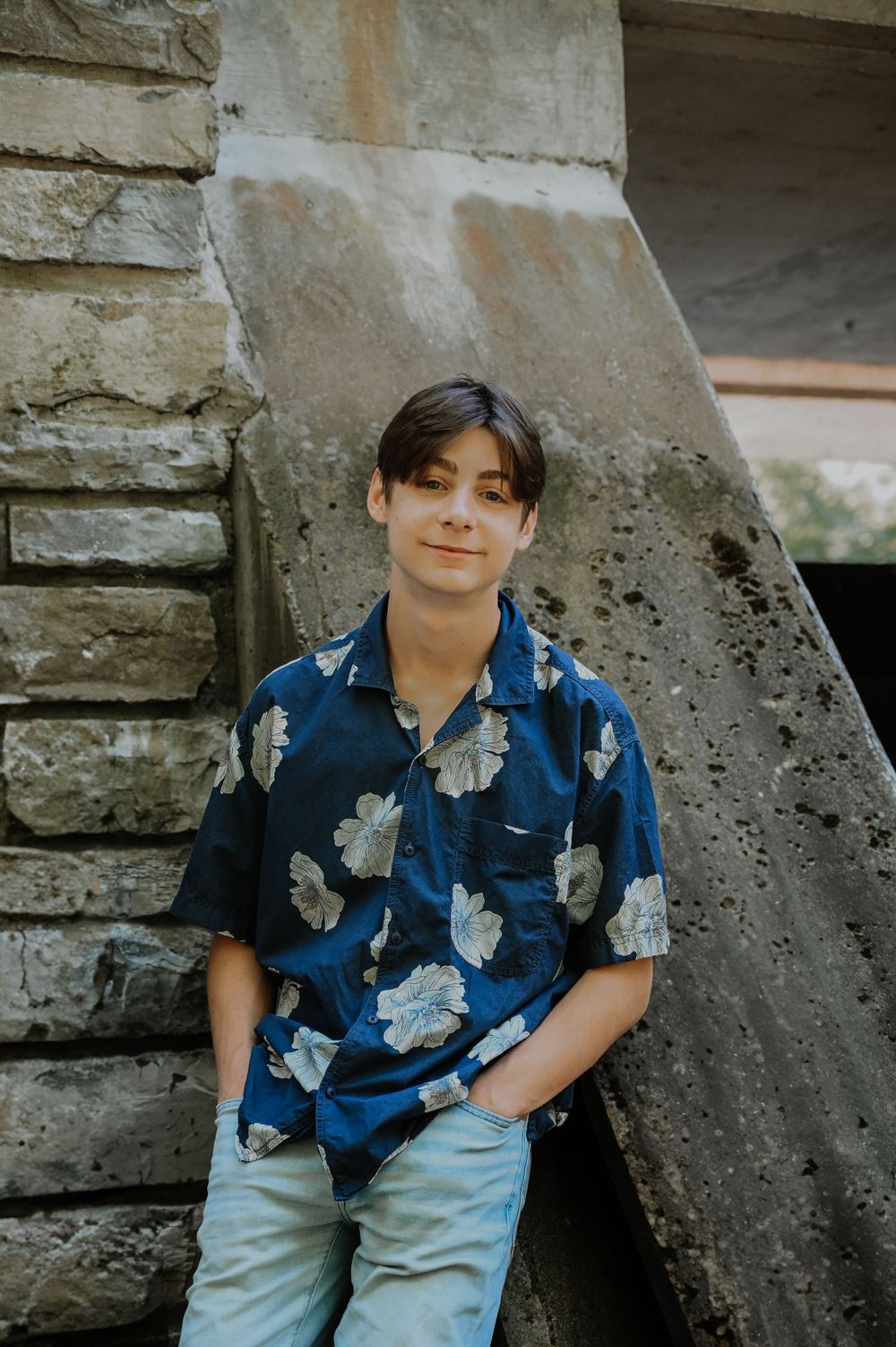Someone wearing a blue floral Hawaiian shirt and light jeans poses casually against a concrete wall structure.
