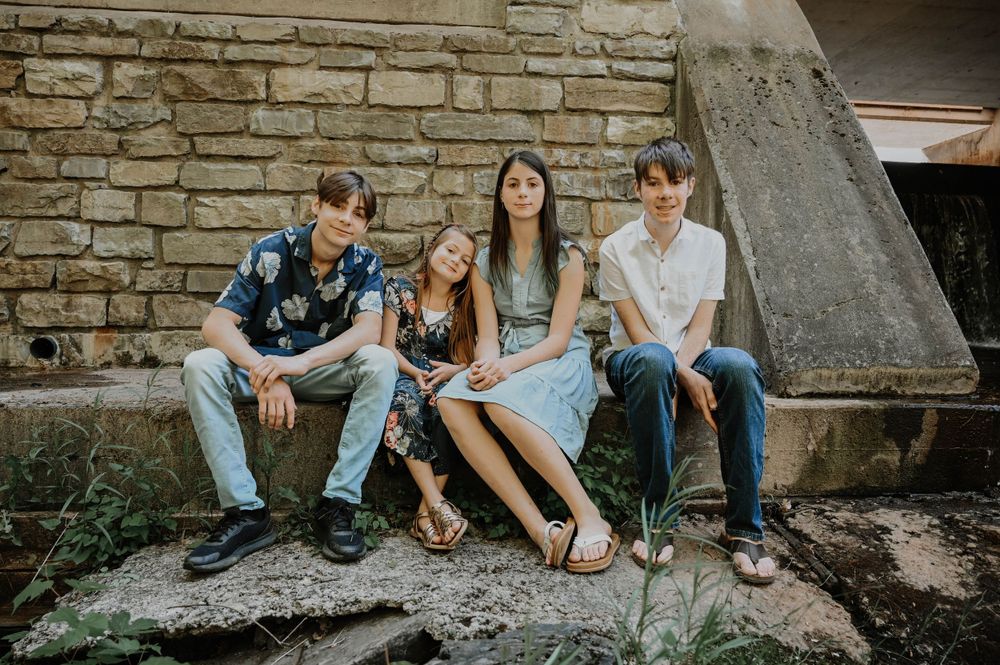 Three young people sit together on stone steps against a weathered brick wall on a sunny day.