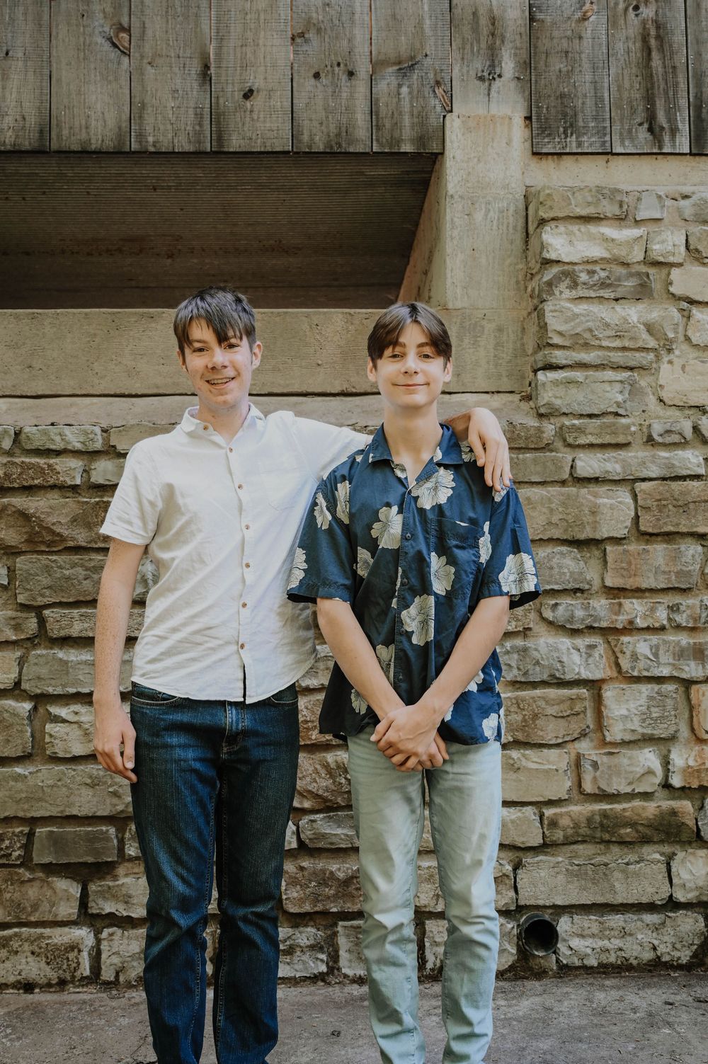 Two young people in casual attire stand against a rustic stone wall and wooden backdrop.