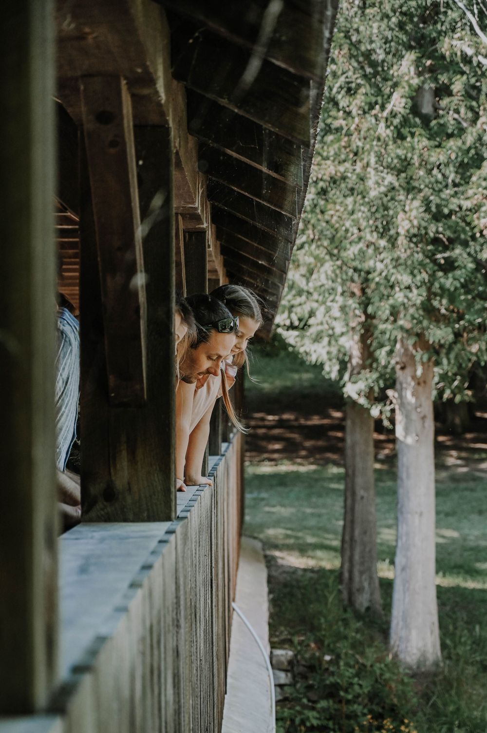 People leaning over wooden railing of covered bridge looking at scenery on sunny summer day.