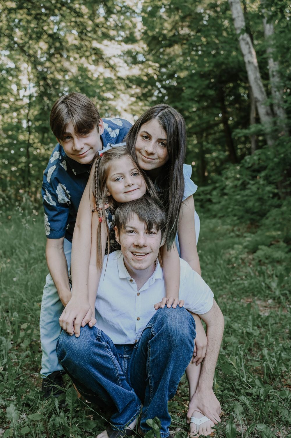 Candid family portrait of siblings and parents sitting together outdoors in a wooded setting.