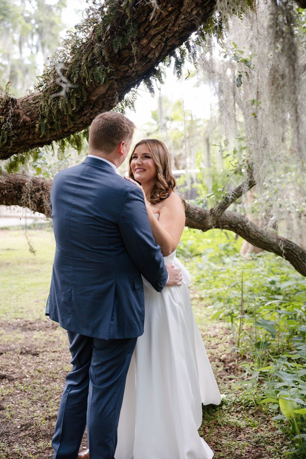 Bride in white gown smiling at groom in navy suit during outdoor first look under a moss-covered oak tree.