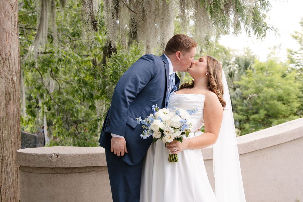 A bride and groom share a romantic kiss outdoors, bride holding white and blue bouquet, groom in navy suit.