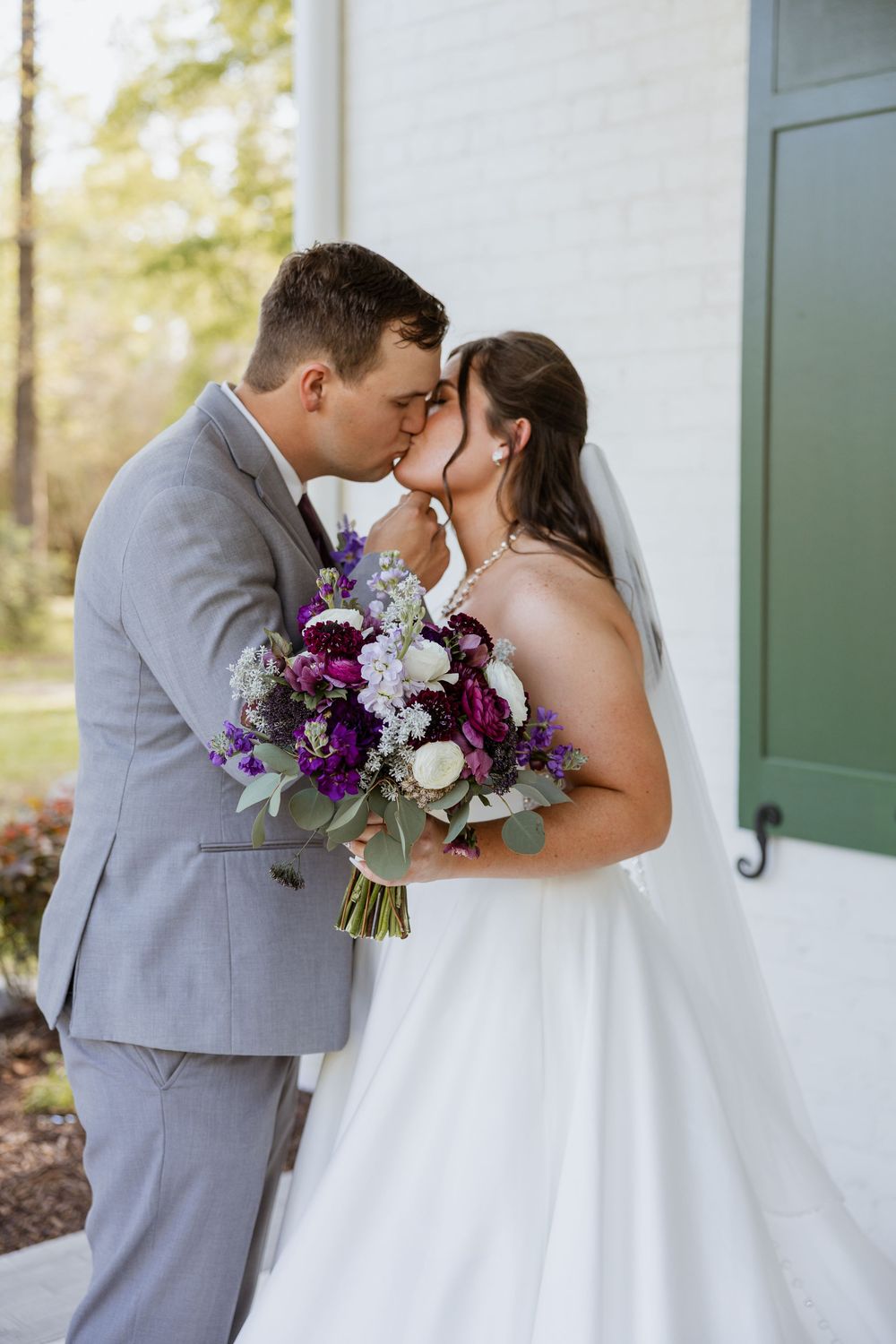 A romantic wedding moment with a beautiful purple and white bouquet, the couple in gray suit and white gown sharing a kiss.