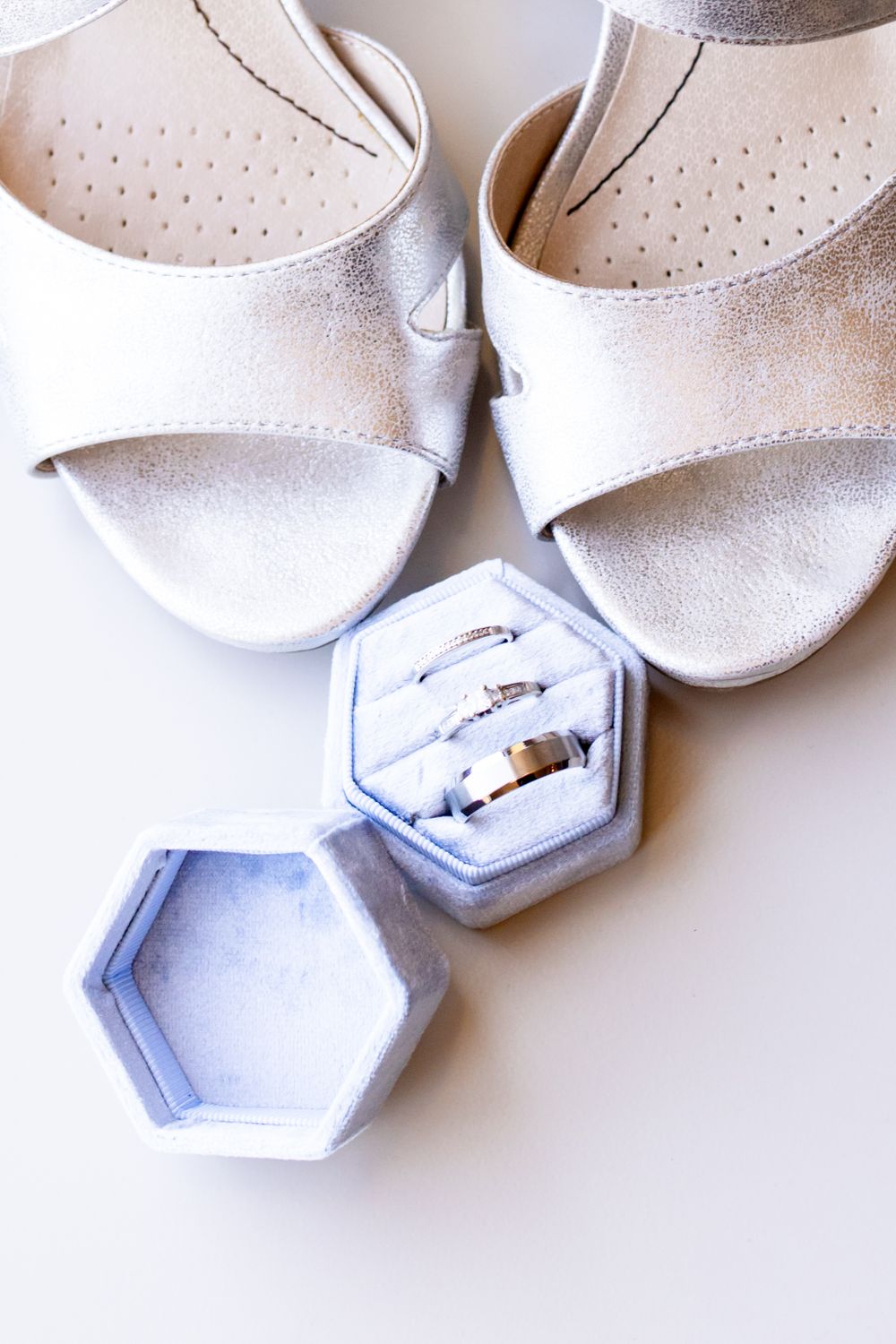 Silver wedding rings in hexagonal velvet box beside elegant metallic heeled sandals on white background.