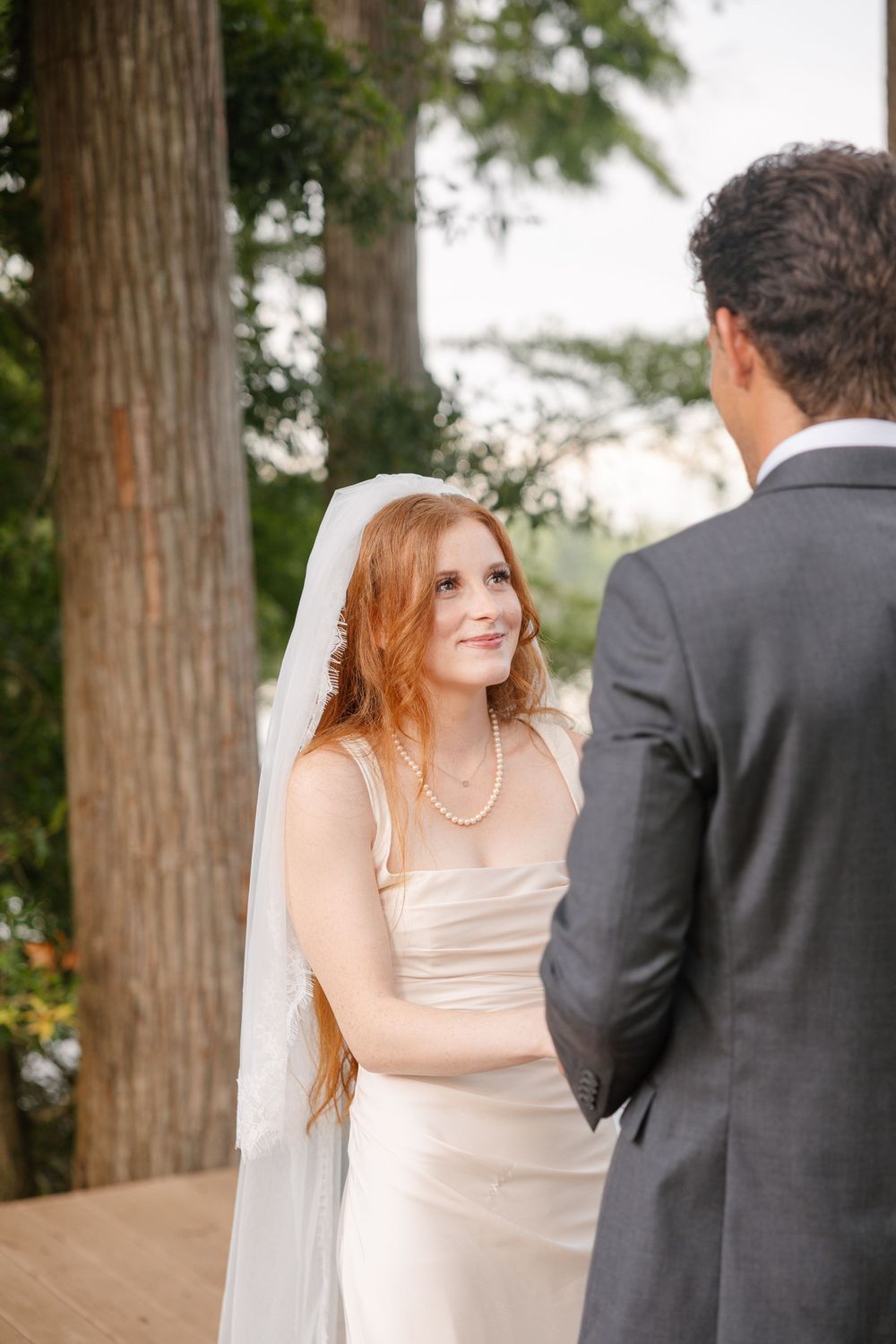 Red-haired bride in white gown and veil smiles at groom in gray suit during outdoor wedding ceremony.