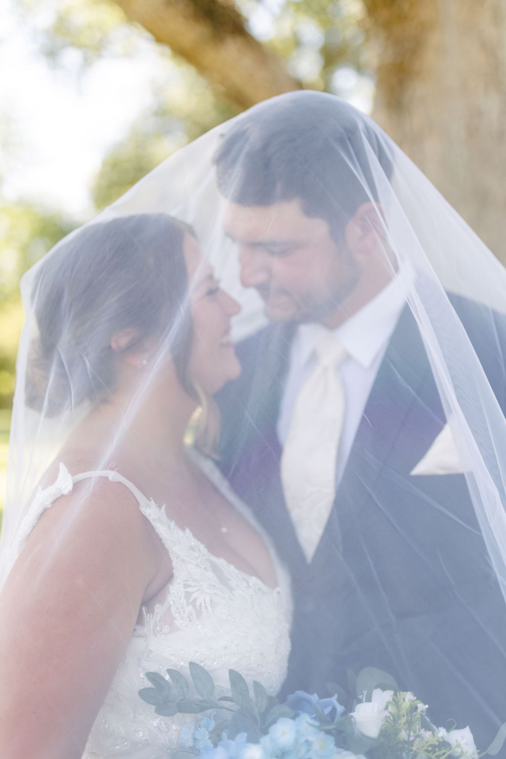 Bride and groom share an intimate moment under a sheer wedding veil, with bride holding blue hydrangea bouquet outdoors.
