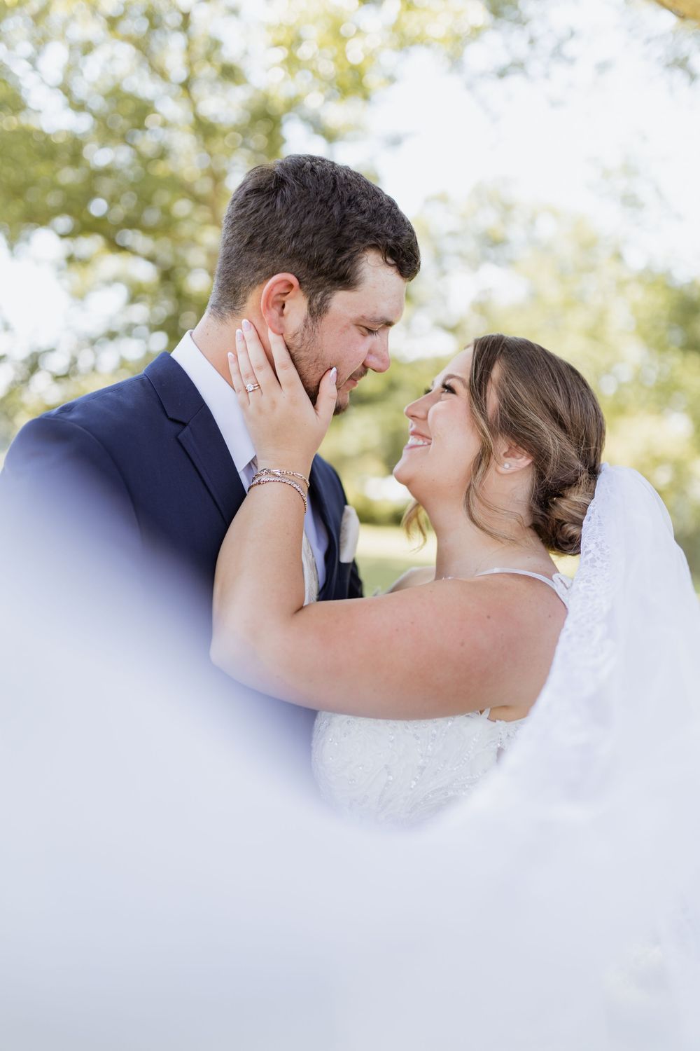 Bride and groom share a romantic moment outdoors, with her hand on his cheek, both smiling, framed by a flowing white veil.