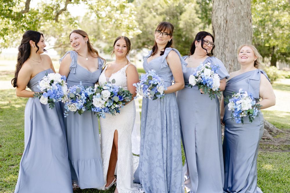 Six bridesmaids in blue dresses holding floral bouquets standing outdoors with a bride in white at a wedding.