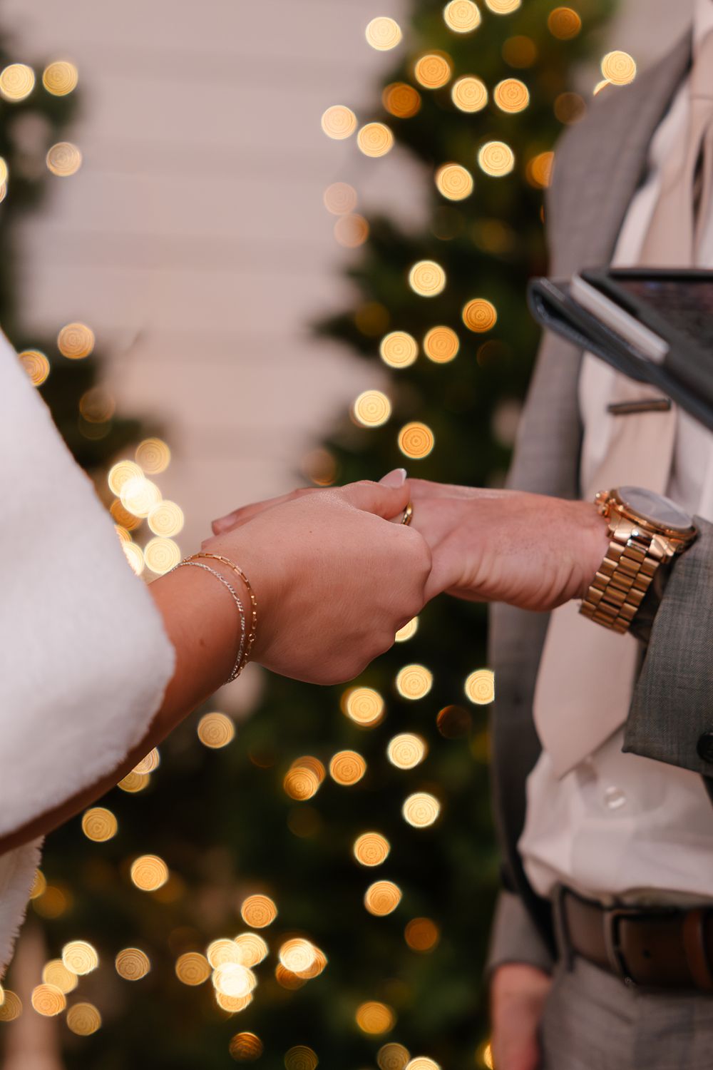Close up of hands exchanging rings with Christmas tree lights twinkling in the background.