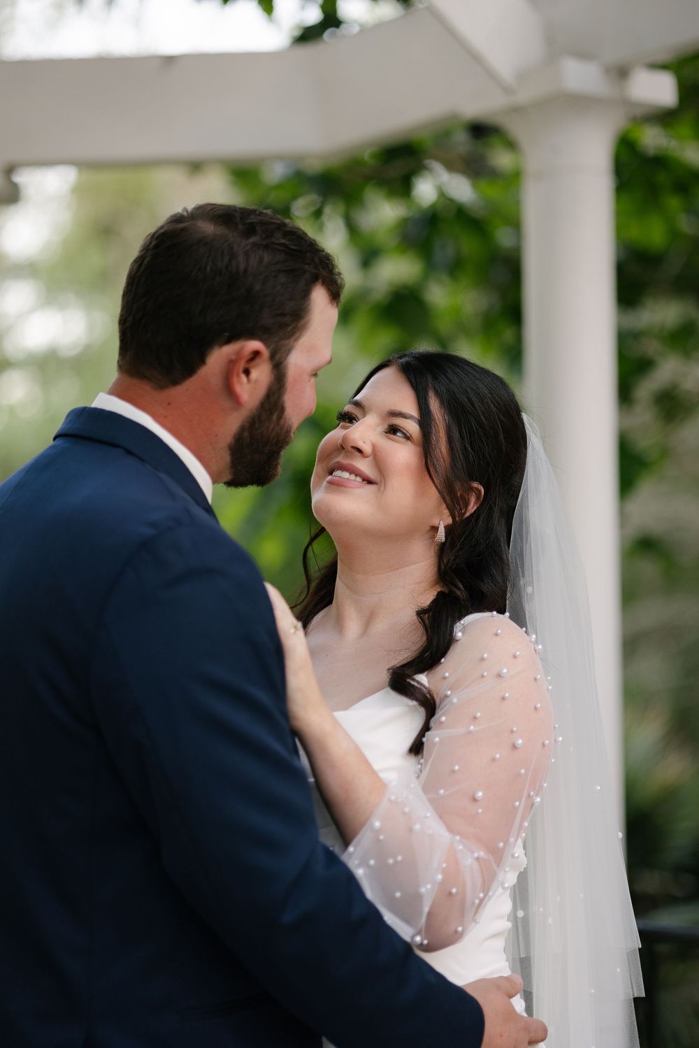 A romantic wedding couple shares an intimate moment between white pillars during their outdoor ceremony.